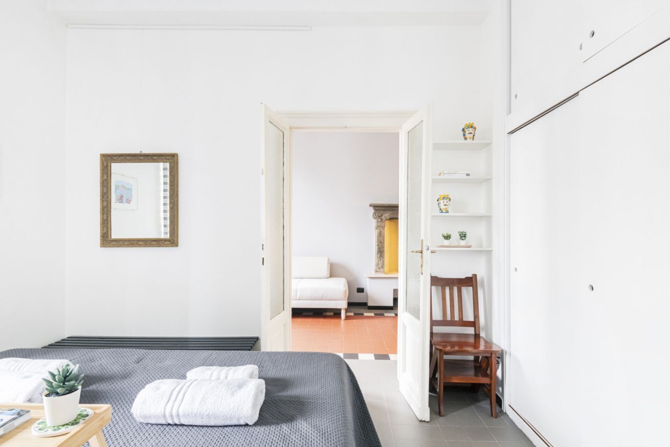 A hotel room with a bed, a chair, a small table with a plant, and a shelf with decorative items.