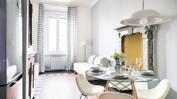 Een eetruimte met een glazen tafel voor vier personen, witte stoelen, een kroonluchter en uitzicht op een raam met gestreepte gordijnen.