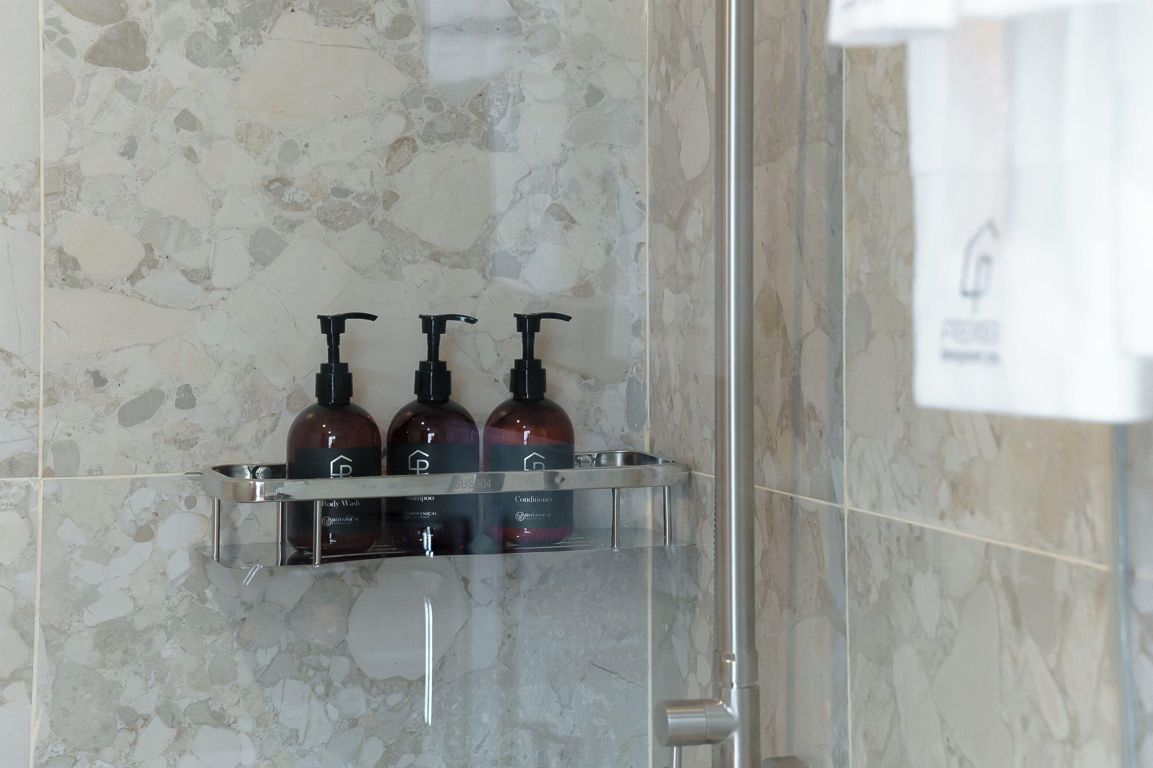 A bathroom with a marble wall and a glass shelf holding three bottles of shampoo and conditioner.