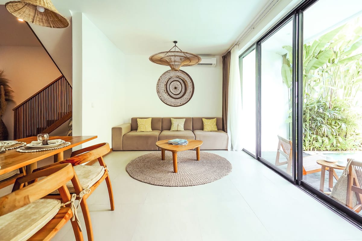 A modern living room with a round coffee table, wooden chairs, a sofa with cushions, a hanging light fixture, and a large window with a view of greenery.