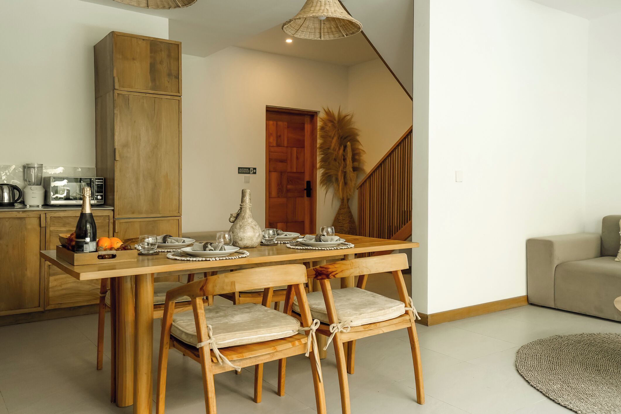 Ein Essbereich mit einem Holztisch, gedeckt für vier Personen, umgeben von Holzstühlen mit gepolsterten Sitzflächen. Im Hintergrund sind eine Holzkommode und eine Treppe zu sehen.