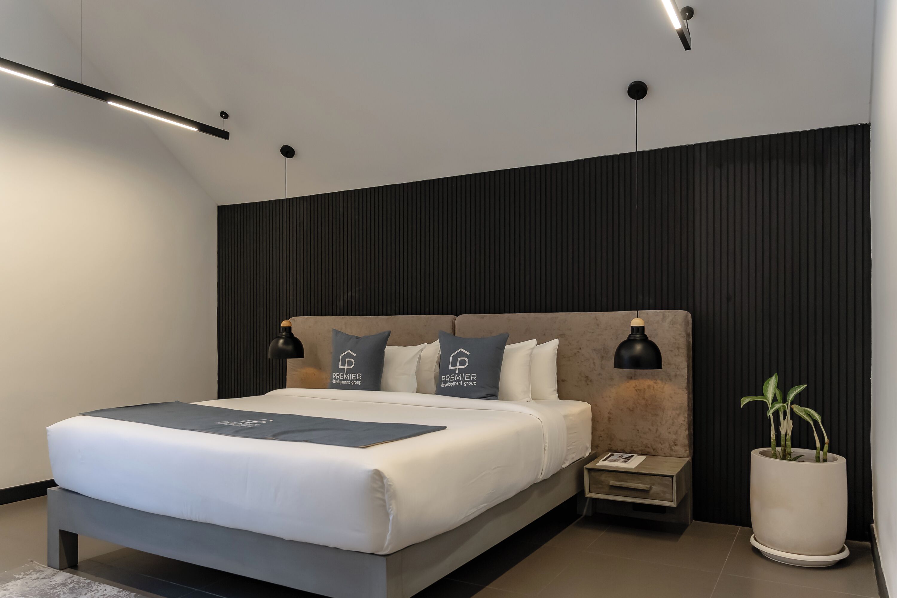A modern bedroom with a large bed, two bedside lamps, a nightstand, a potted plant, and a wall-mounted light fixture.