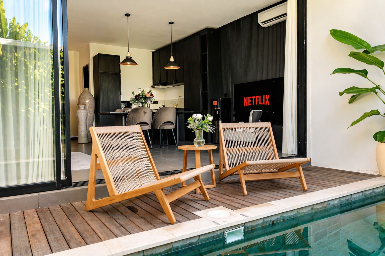 Zona de piscina amb dues cadires de vimet, una taula petita i vista a una cuina i zona de menjador modernes.