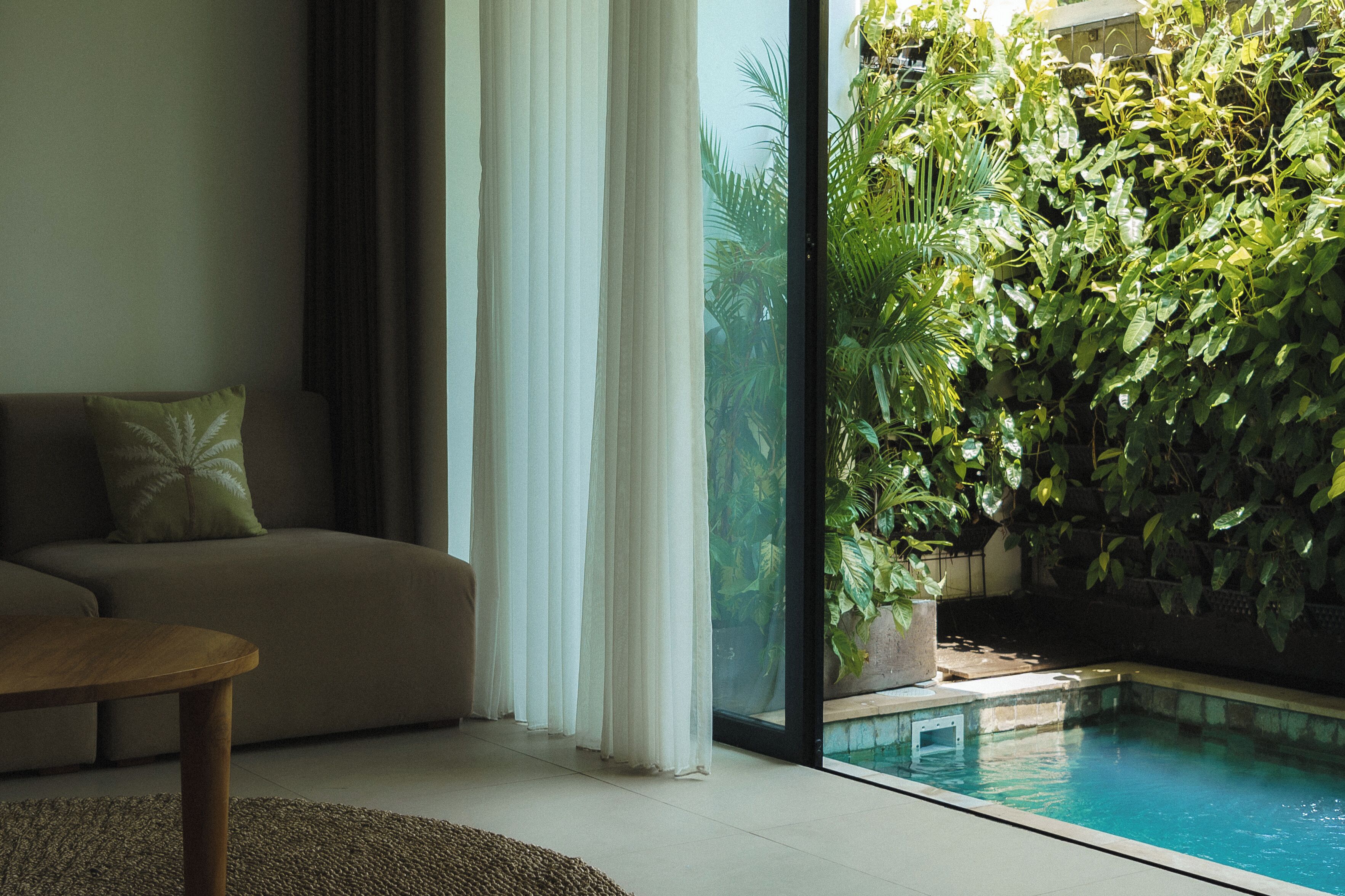 Una sala de estar con sofá, una mesita de centro y vistas a una piscina y exuberante vegetación.