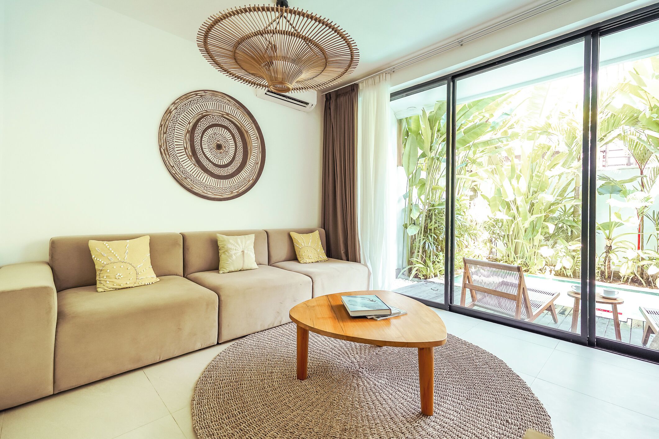 A modern living room with a beige sofa, a round wooden coffee table, and a large window with a view of tropical plants.