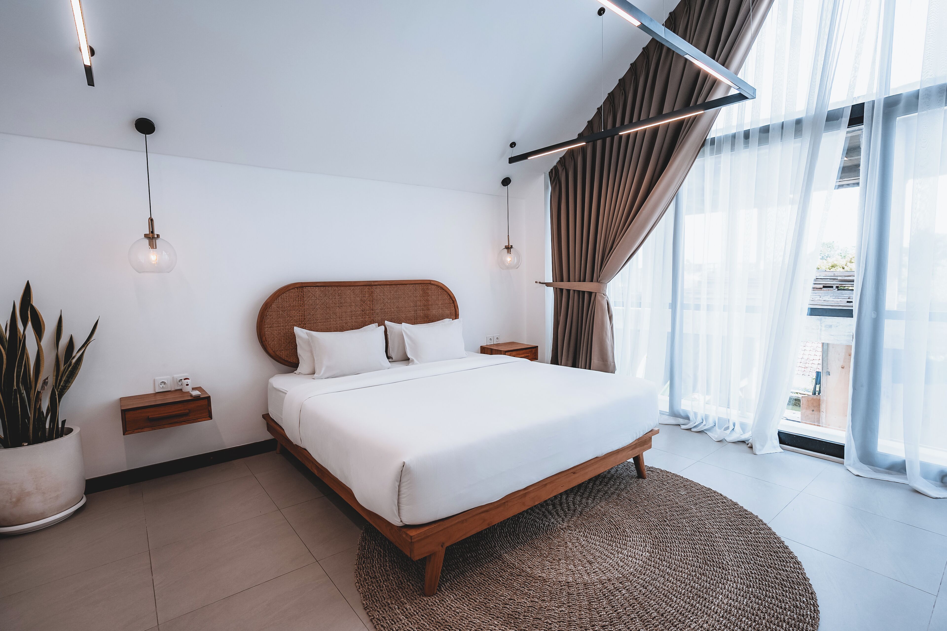 A modern bedroom with a wooden bed, bedside tables, a potted plant, and sheer curtains.
