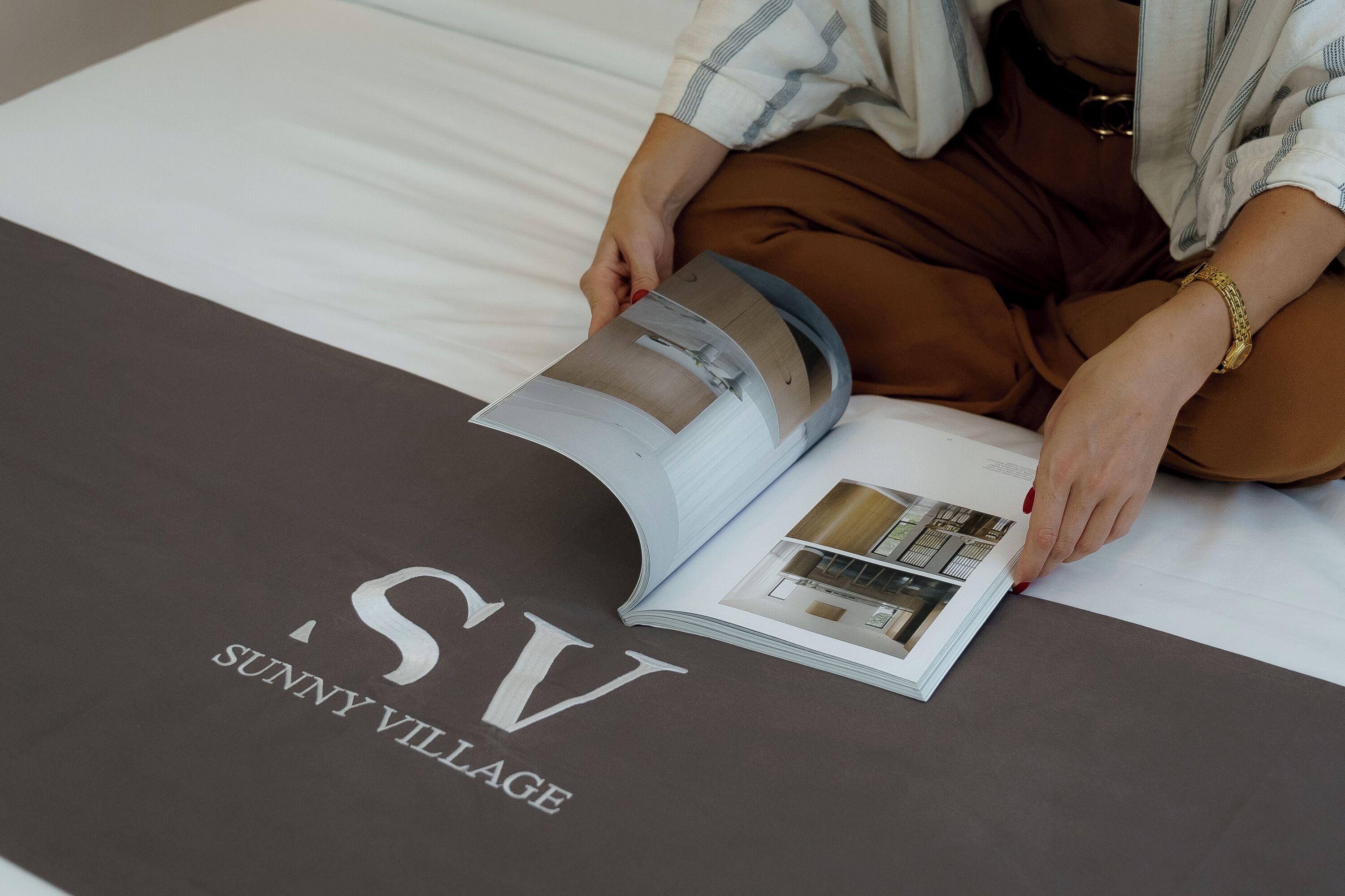 A person reading a magazine on a bed with a bedspread that has 'SUNNY VILLAGE' printed on it.