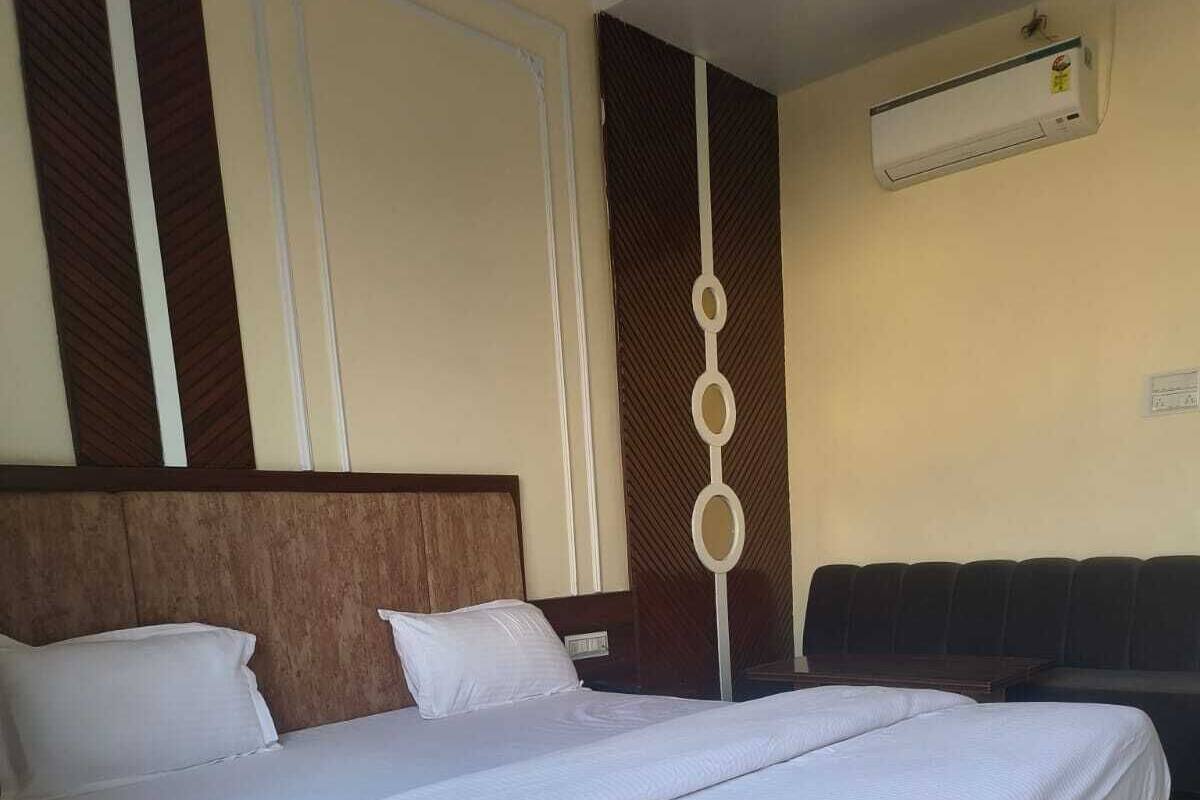 A hotel room with a large bed, a wooden headboard, a wall-mounted air conditioner, and a dark-colored sofa.