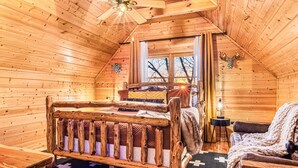 A log cabin bedroom with a wooden bed, a chair, a small table, and a window with curtains.