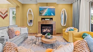 A living room with a fireplace, a sofa, a yellow armchair, and a television displaying a beach scene. - Destin: Private Beach, Pool, 4 En-Suites (Destin)