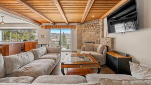 A modern living room with a stone fireplace, wooden ceiling beams, and a large window overlooking a scenic landscape. - Estate on Bow River 25 Min to  DT YYC 5BR w AC (Calgary)