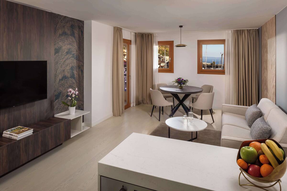 Ein modernes Wohnzimmer mit Flachbildfernseher, einem Essbereich mit rundem Tisch und Stühlen, einem weißen Sofa und einer Obstschale.