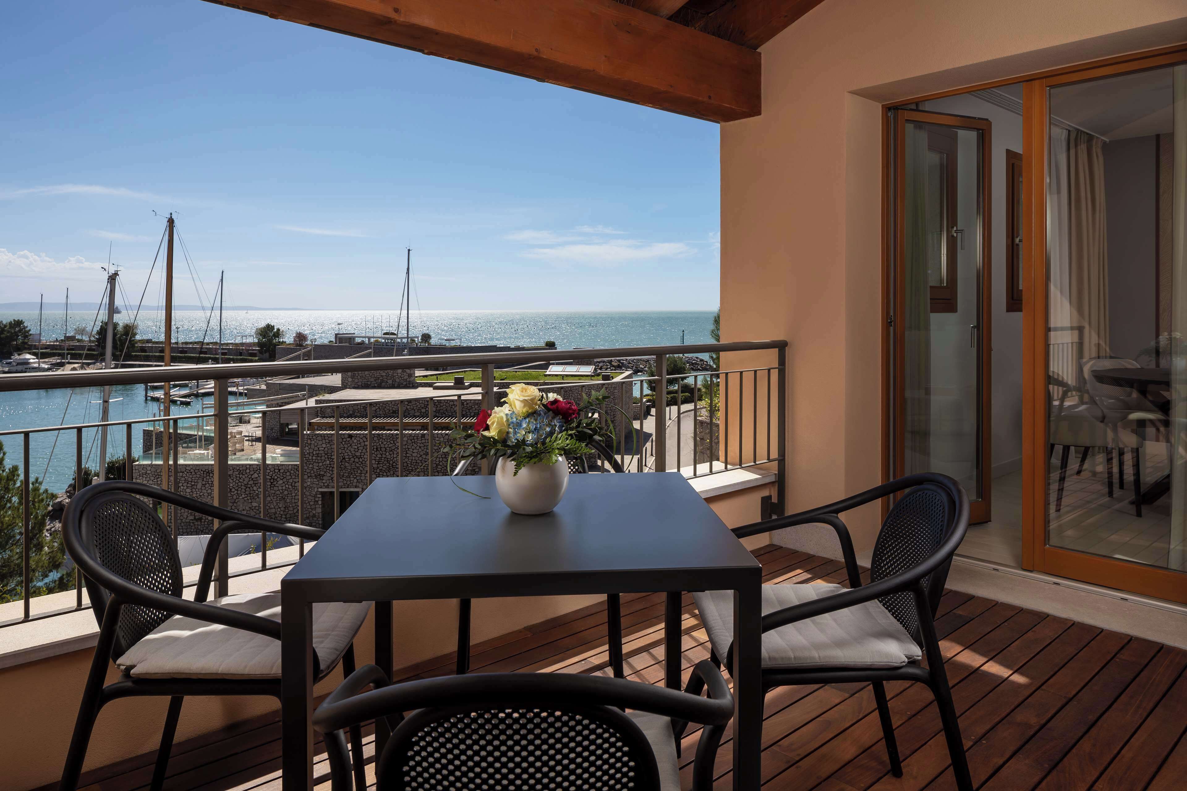 Un balcone con tavolo e sedie, un vaso di fiori e vista sul mare e sulle barche.