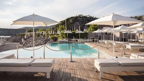Un espace aménagé au bord de la piscine, avec des chaises longues et des parasols, une terrasse en bois et une vue sur la mer et les collines en arrière-plan.