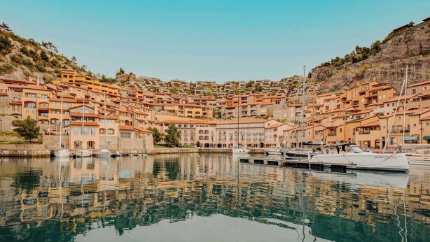 Une ville côtière avec des bâtiments à étages le long du front de mer, un port de plaisance rempli de bateaux et un reflet limpide dans l’eau.