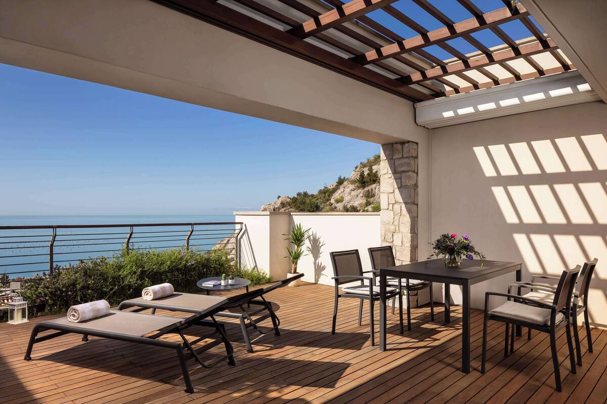 Eine Holzterrasse mit Tisch und Stühlen, einer Pergola und Blick auf das Meer.