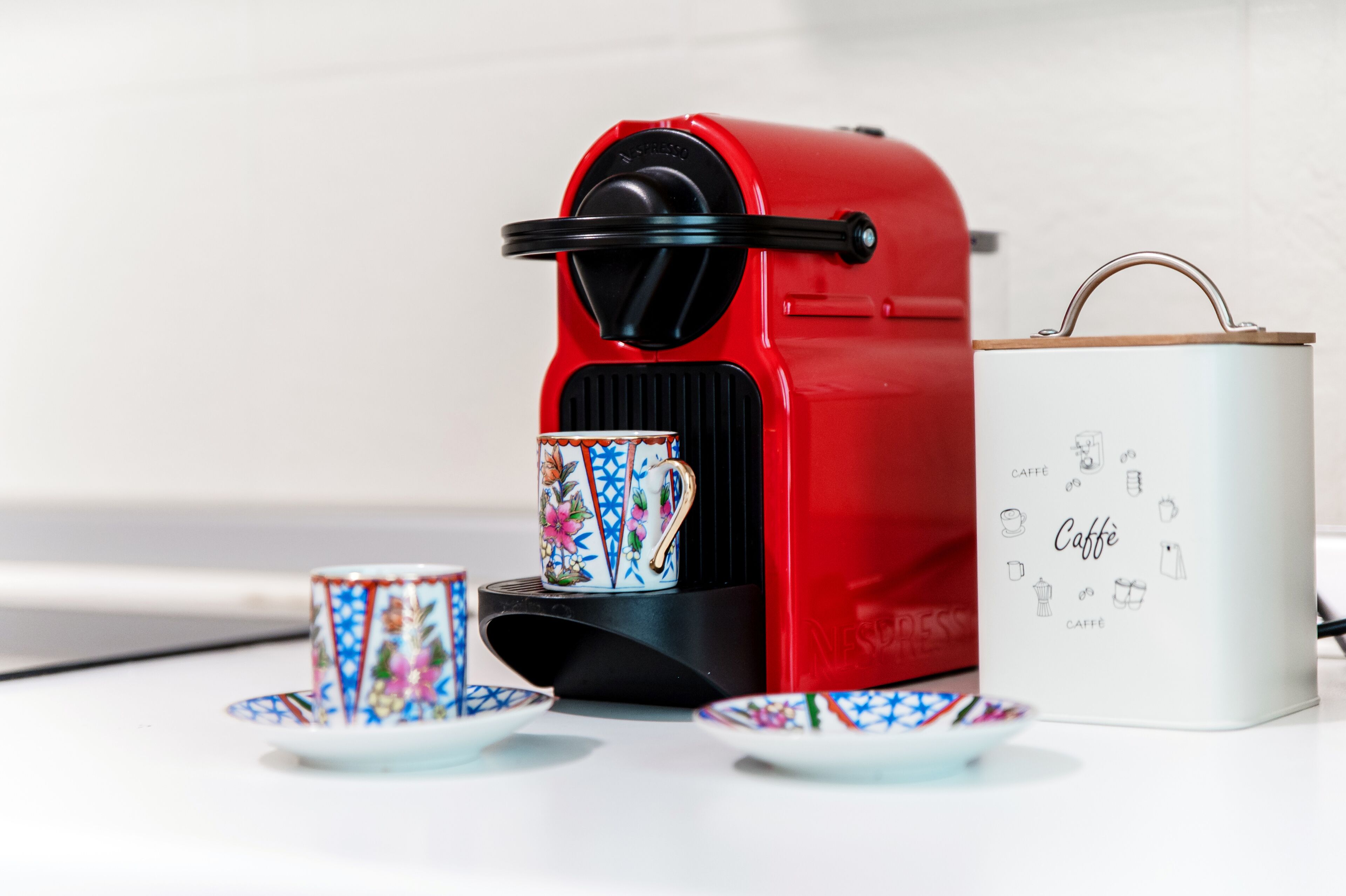 A red coffee maker with a coffee pod and matching cups, a white box with coffee-related illustrations, and a handle on the box.