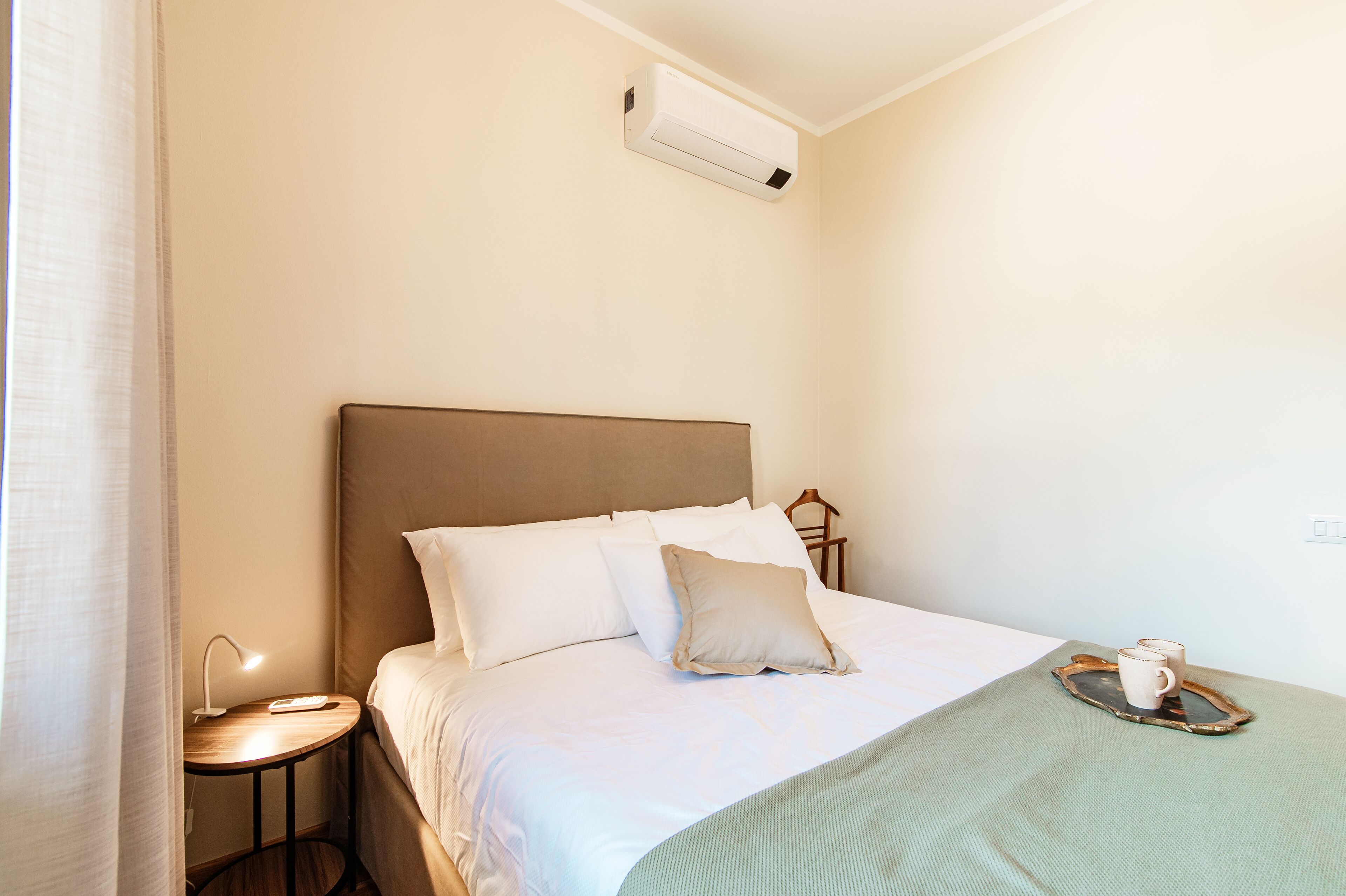 A neatly made bed with white linens and a beige pillow, a bedside table with a lamp, and a tray with two mugs on the bed.