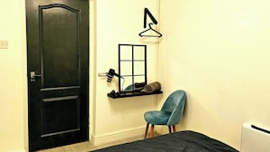 Una habitación con una puerta negra, un estante pequeño con un espejo y una toalla, una silla azul y un radiador.