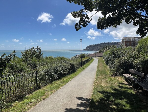 A coastal path with a metal fence, a bench, and a building in the background.