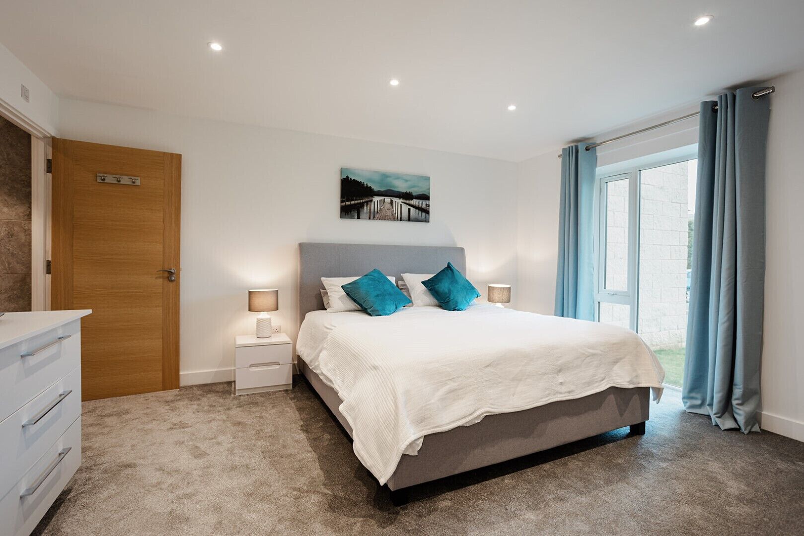 A bedroom with a bed, bedside tables, a nightstand, a dresser, a window with curtains, and a door.