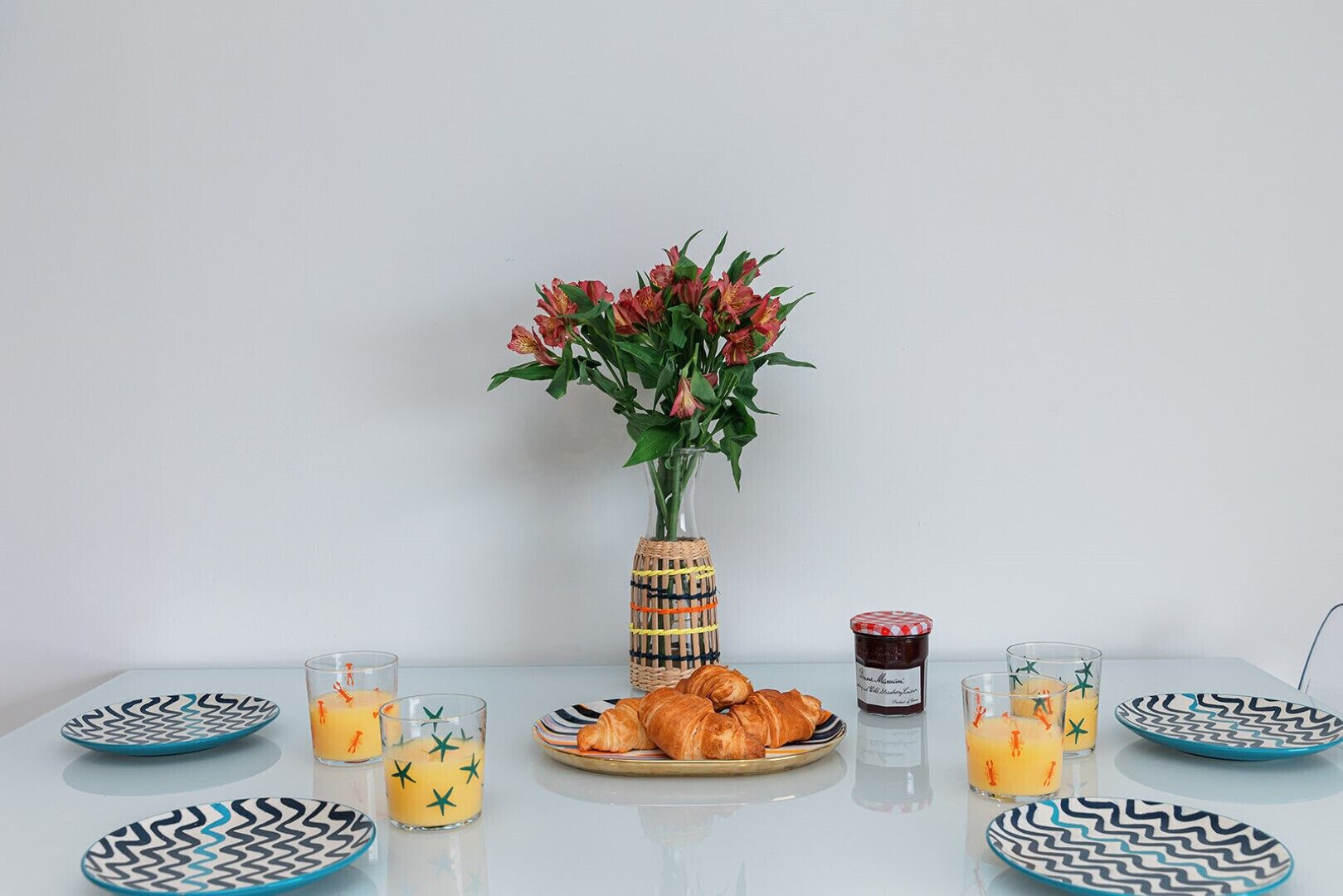 En frukostuppsättning med croissanter, apelsinjuice och ett glas marmelad på ett bord med mönstrade tallrikar.