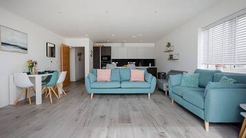Un salon moderne avec un canapé de couleur turquoise, un coin repas avec des chaises blanches et une cuisine en arrière-plan.