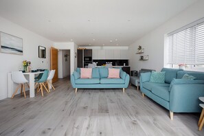 Un salon moderne avec un canapé de couleur turquoise, un coin repas avec des chaises blanches et une cuisine en arrière-plan.