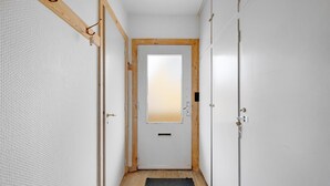 Un couloir étroit, avec une porte en bois au bout, bordé de murs blancs et surmonté d’une moquette grise.