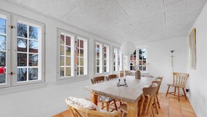 Un coin repas avec une grande table en bois et des chaises en osier, de grandes fenêtres à cadres blancs et un sol carrelé.