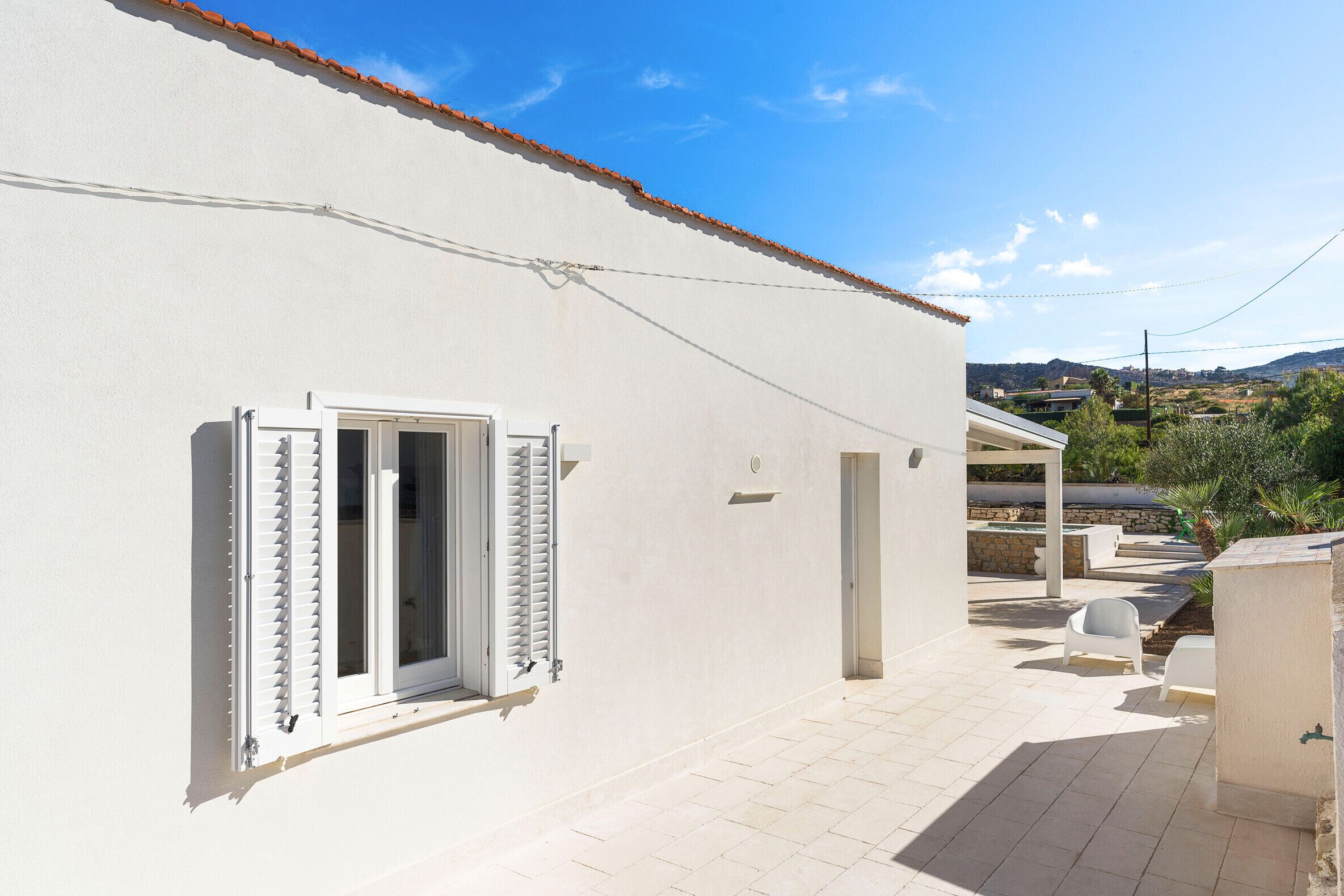 A white building with a tiled patio, a window with shutters, and a view of a hilly landscape.