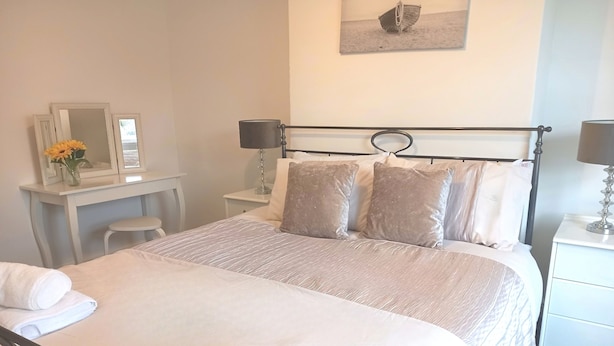 Una cama bien hecha con almohadas, una mesita de noche con lámpara, un escritorio pequeño con espejo y florero, y un cuadro colgado en la pared.