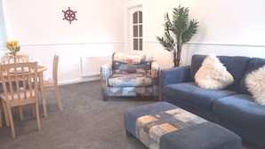 Un salón con un sofá azul, un sillón estampado, una mesa de comedor de madera y sillas, y un radiador.