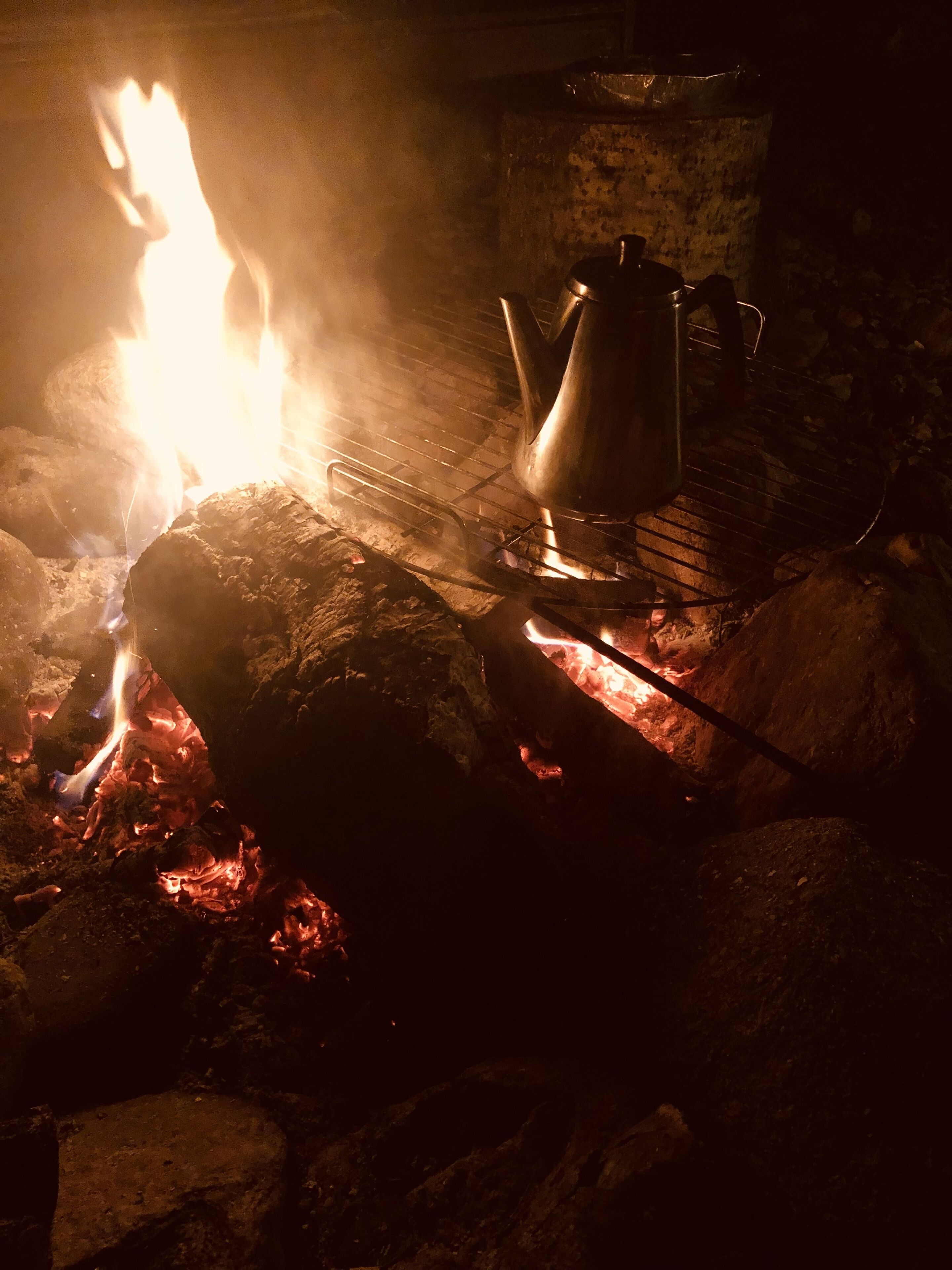 Un coin cuisine rustique aménagé à l’extérieur, avec une marmite en métal et un pichet en métal posés sur une grille métallique au-dessus d’un lit de braises incandescentes.