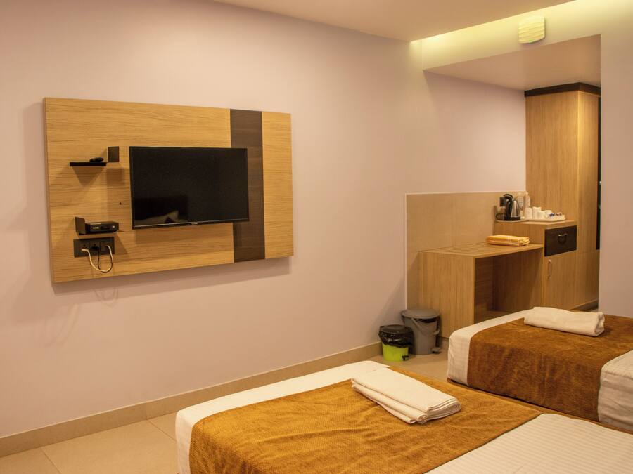 A hotel room with two beds, a TV mounted on the wall, a mirror, and a small kitchenette area.