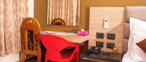 A hotel room with a wooden desk, a red chair, a bed with white bedding, a bedside table with a phone and a bottle, and a wall-mounted control panel.
