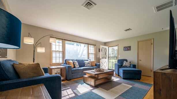 A spacious living room with a blue sofa, a wooden coffee table, and a blue armchair. There are large windows allowing natural light and a view of the outdoors. - Grandma's House | Classic Indy Charm, Private Yard (Indianapolis)