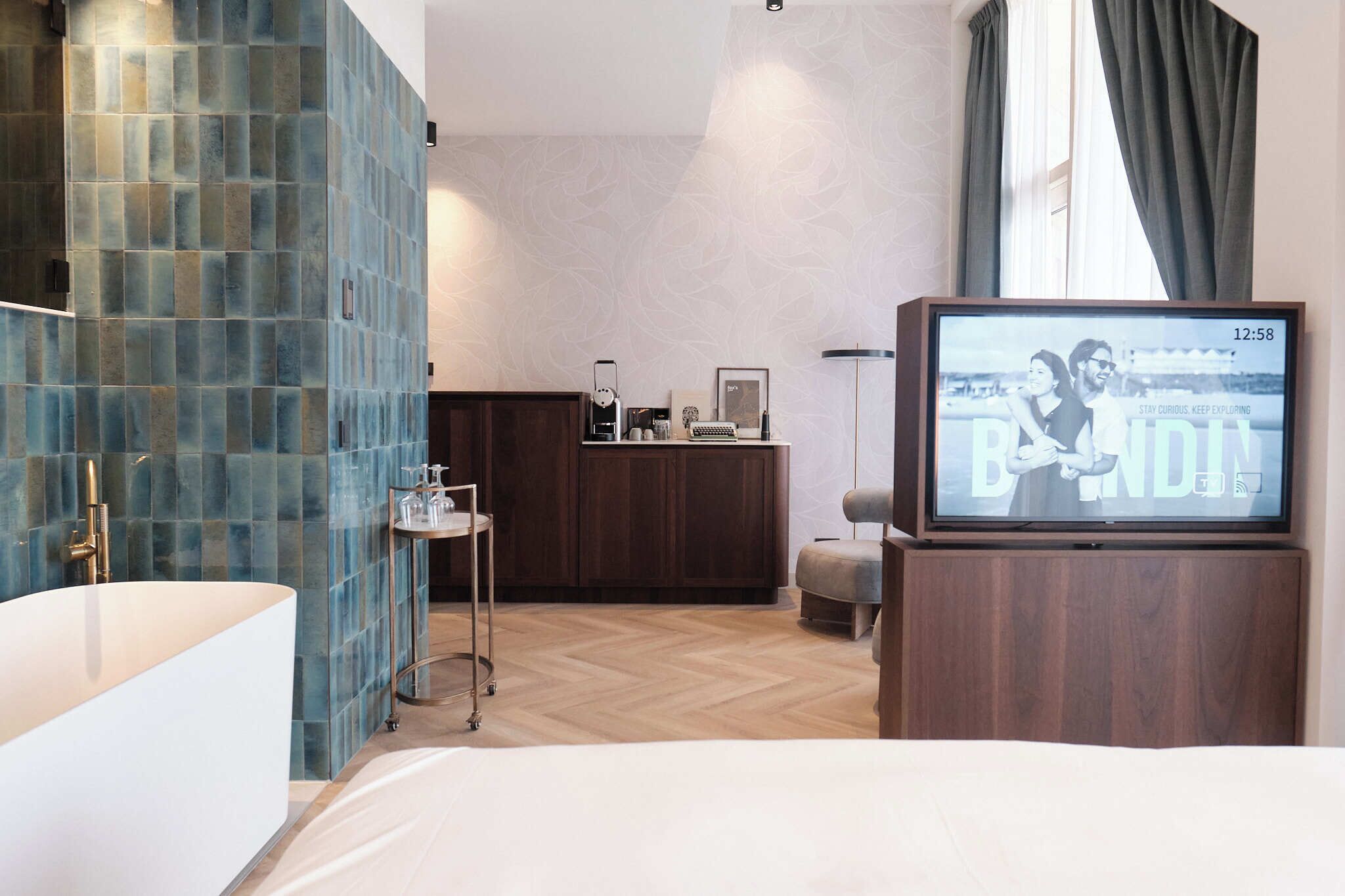 Een moderne hotelkamer met een groot bed, een televisietoestel waarop een film draait, een houten kast en een badkamer met een blauwe betegelde wand.