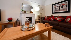 A living room with a wooden coffee table, a fireplace, a brown leather sofa with red cushions, a framed picture on the wall, and a tall floor lamp. - Ladyland Cottage in Maybole (Maybole)