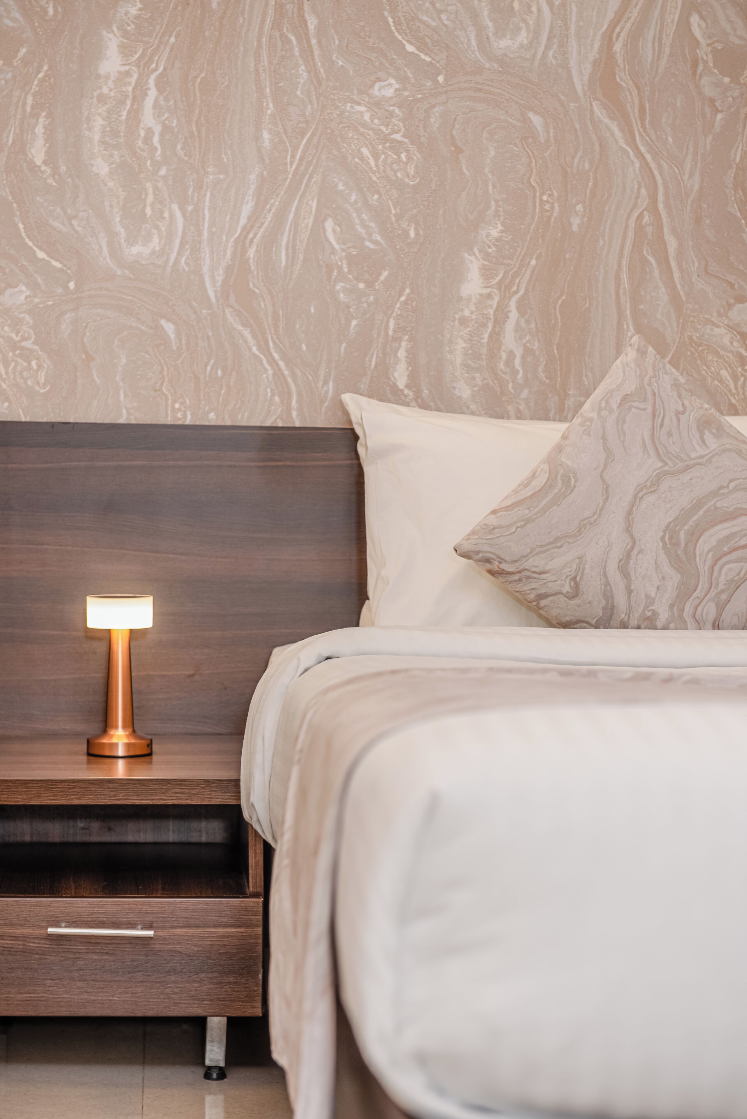 A bedroom with a wooden nightstand, a bedside lamp, a bed with white bedding, and a marble-patterned wall.