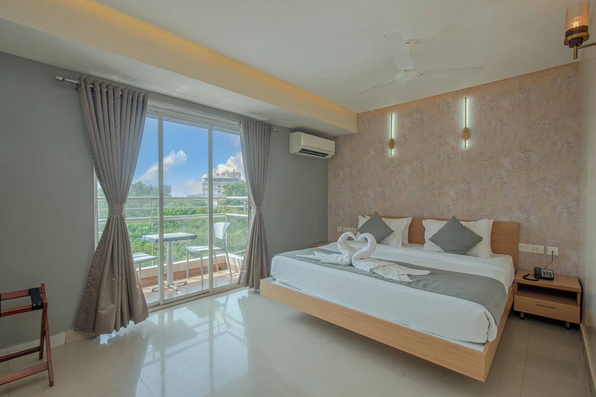 A modern hotel room with a large bed, a balcony with a table and chairs, and a view of the cityscape.