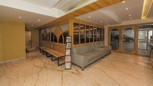 A lobby with a wooden bench, a grey sofa, and a magazine rack. - Hotel Shoolin Grand (Mangaluru)