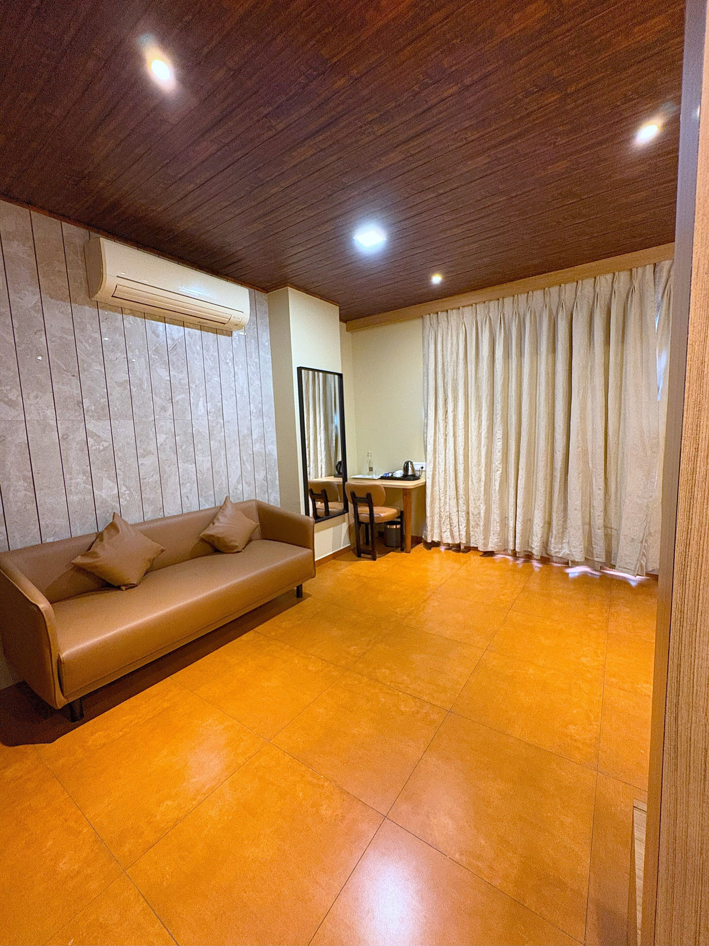 Una habitación con techo de madera, un sofá color beige con almohadas, una pequeña mesa con sillas y un gran ventanal con cortinas.