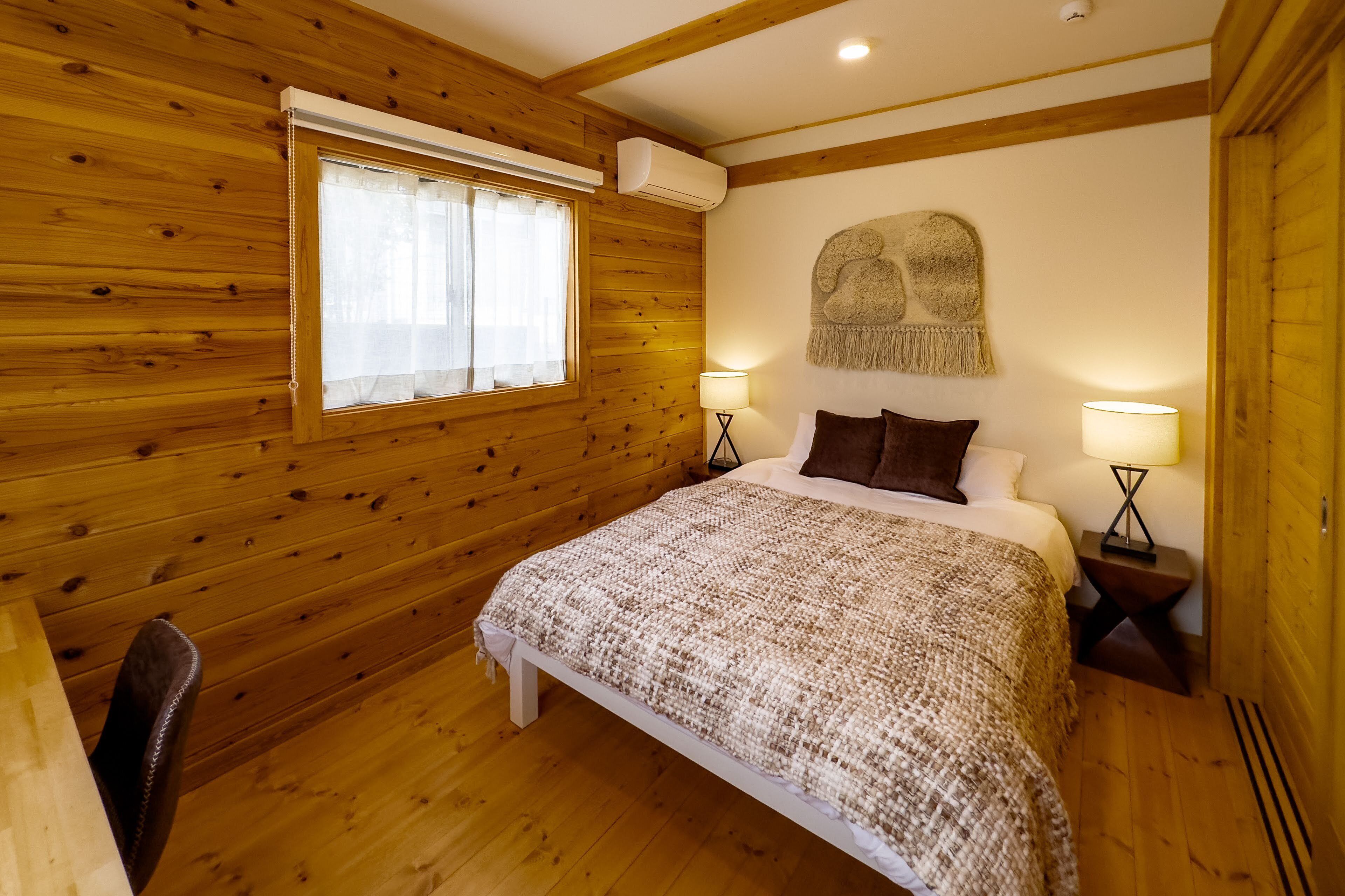 Quarto com painéis de madeira, uma cama, duas luminárias de cabeceira, uma janela com cortinas e um objeto decorativo fixado na parede.