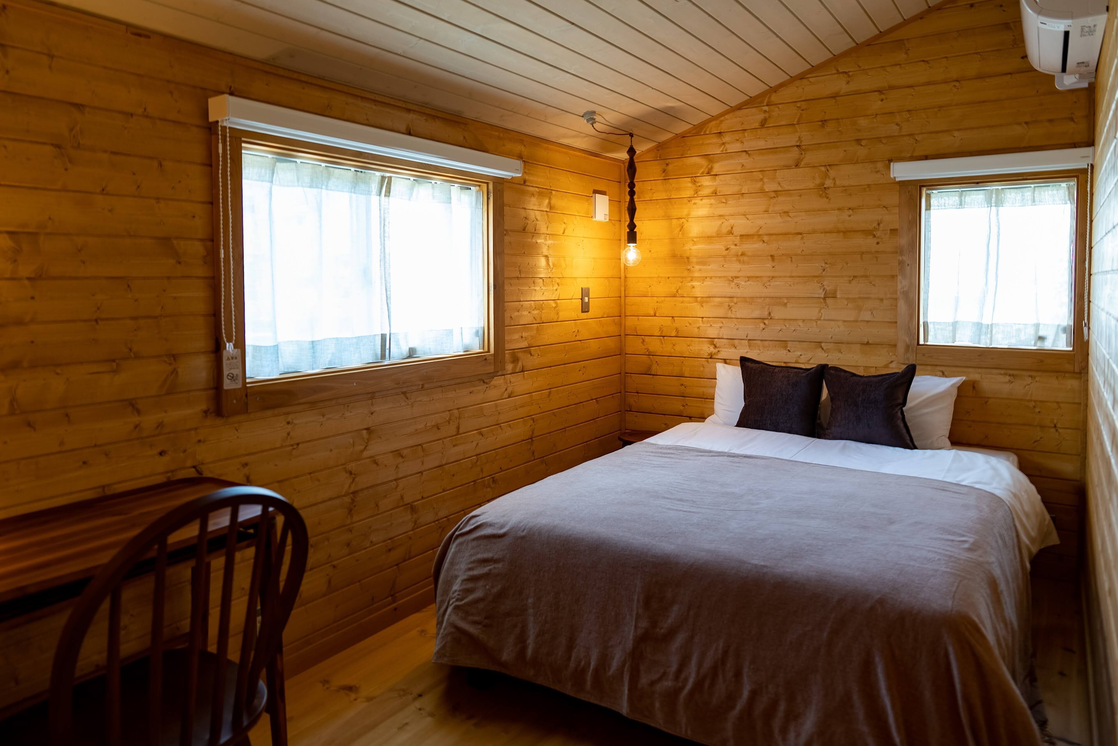 A wooden cabin room with a bed, two windows, a desk, and a chair.