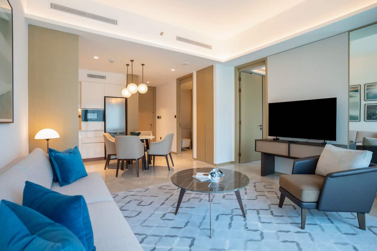A modern living room with a sofa, armchairs, a coffee table, and a TV. There is a dining area with chairs and a table, and a kitchen in the background.