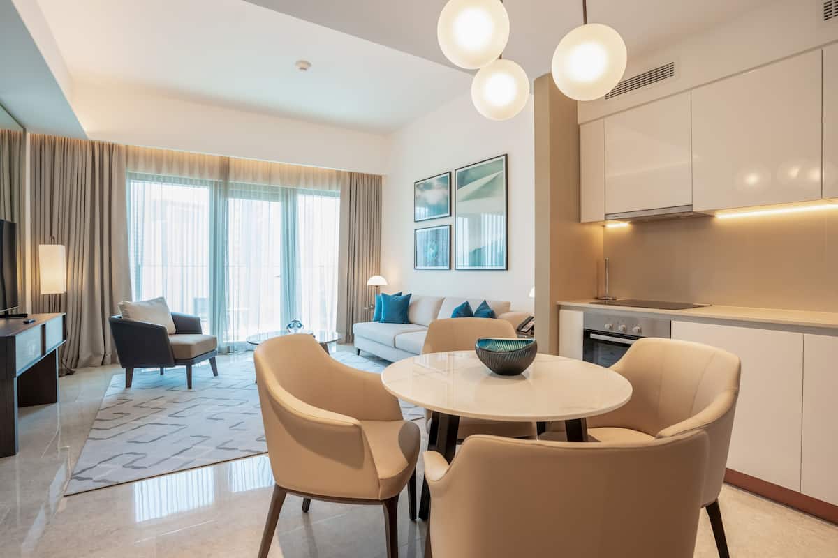 A modern hotel room with a dining area, a sofa set, a TV, and a kitchen area.