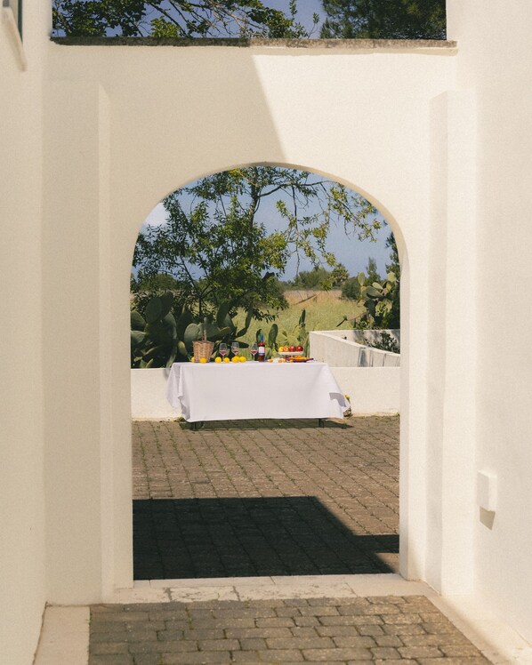 An outdoor dining area with a table set under a white archway, surrounded by greenery and a clear sky. - Tenuta Doxi (Sannicola)