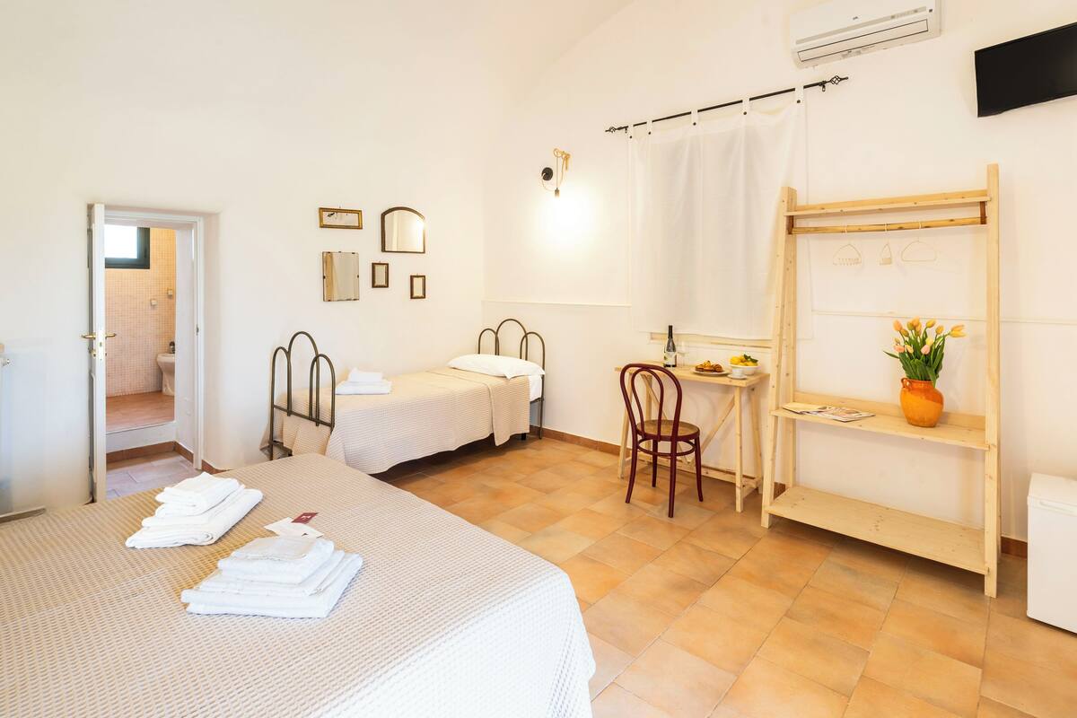 A hotel room with two single beds, a desk with a chair, a small table with a vase of flowers, and a wooden shelf.