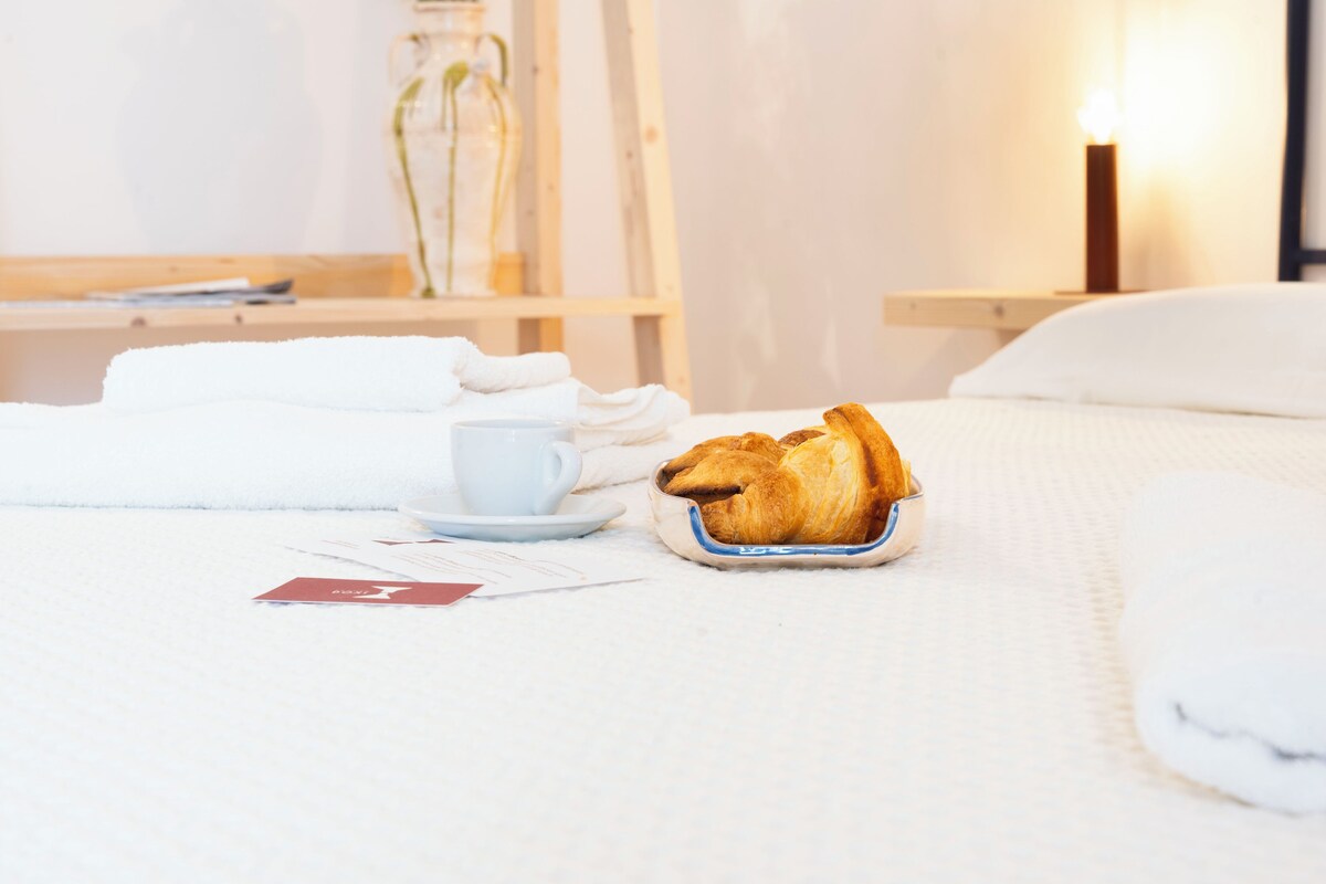A neatly made bed with a white duvet, a white pillow, and a small tray with a croissant. A white cup and saucer are also visible on the bed.